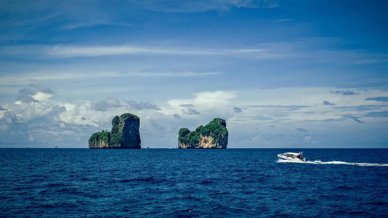 A white speedboat crossing a deep blue sea, two jungle-covered limestone islets on the horizon