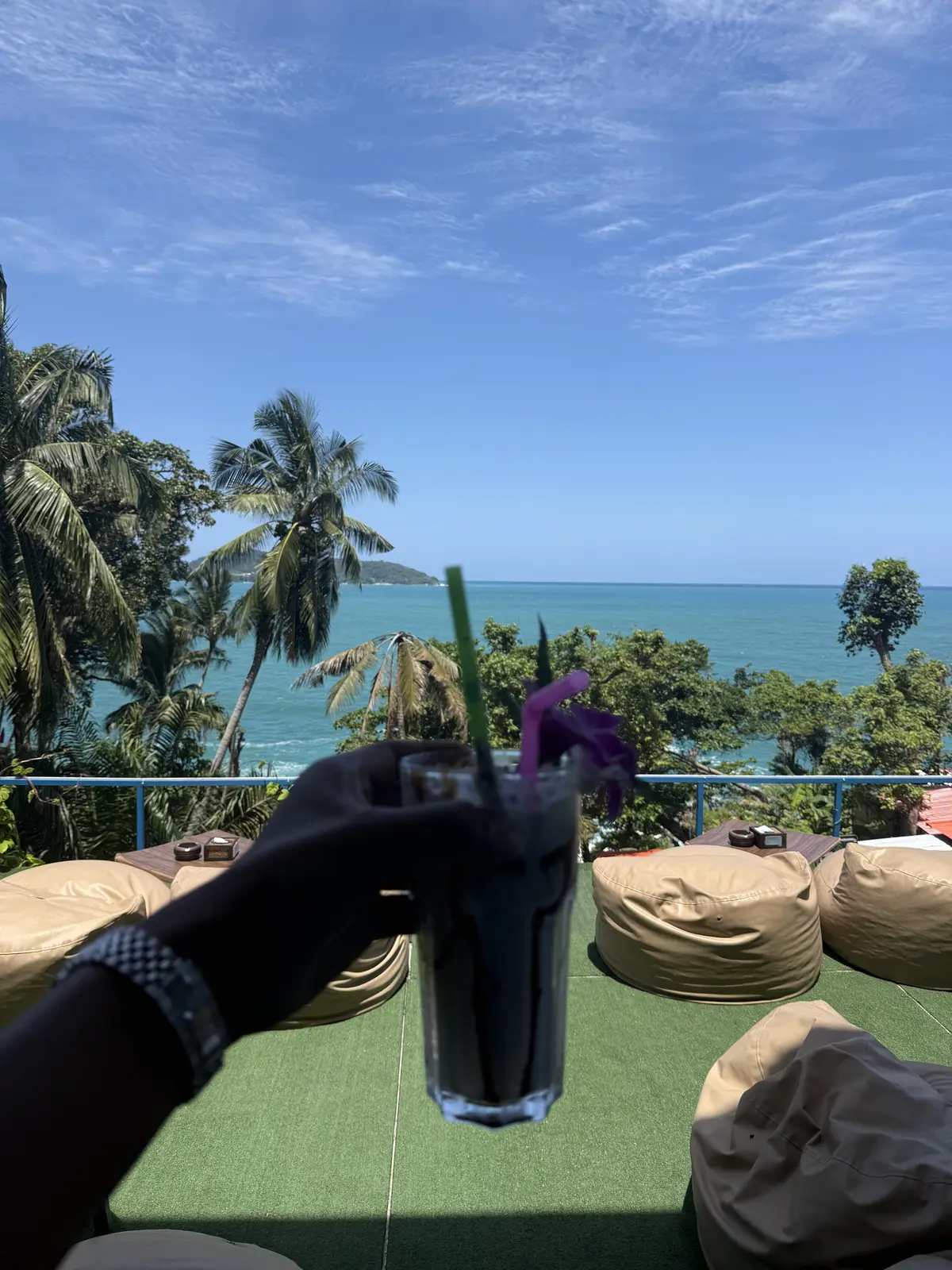 Verre tenu à la main devant des poufs et la mer en fin de journée, dans un bar de plage de Phuket