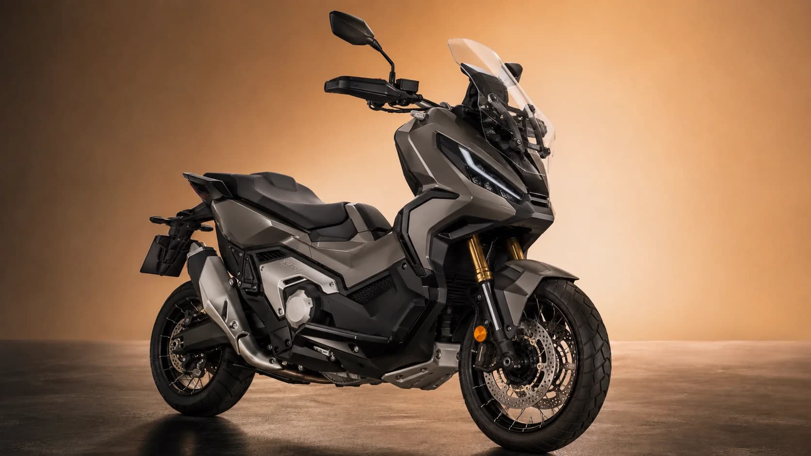 Honda X-ADV 750 gris titane vu de trois quarts avant, sur fond ambré