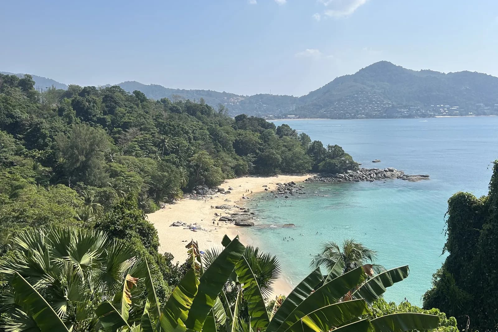 Vue plongeante depuis la colline sur une plage de sable clair de la côte ouest de Phuket : eau turquoise, rochers, jungle au premier plan et collines habitées au fond