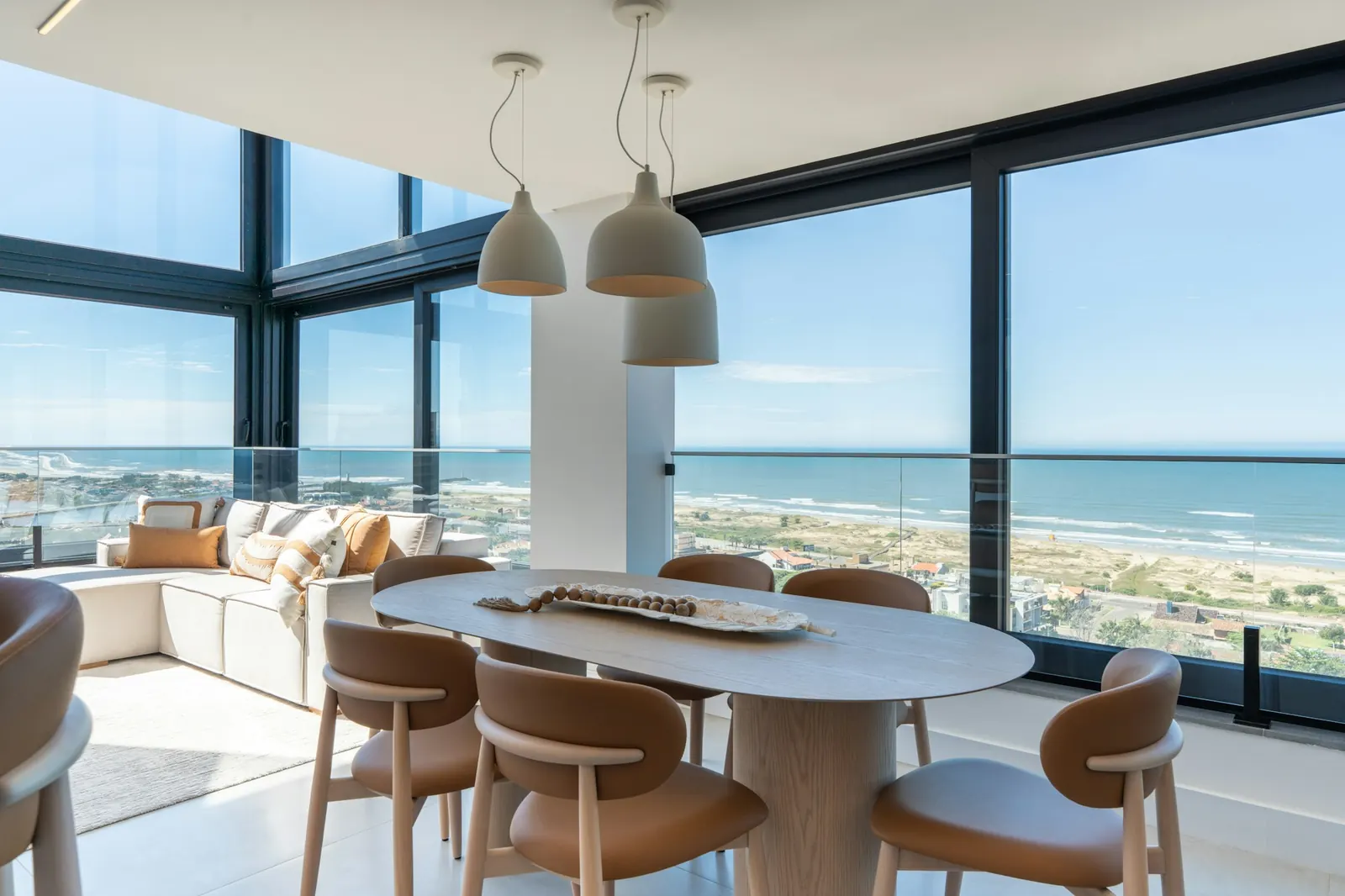 Bright living and dining room in an upper-floor apartment: oval table, light sofa and floor-to-ceiling windows onto the beach and sea