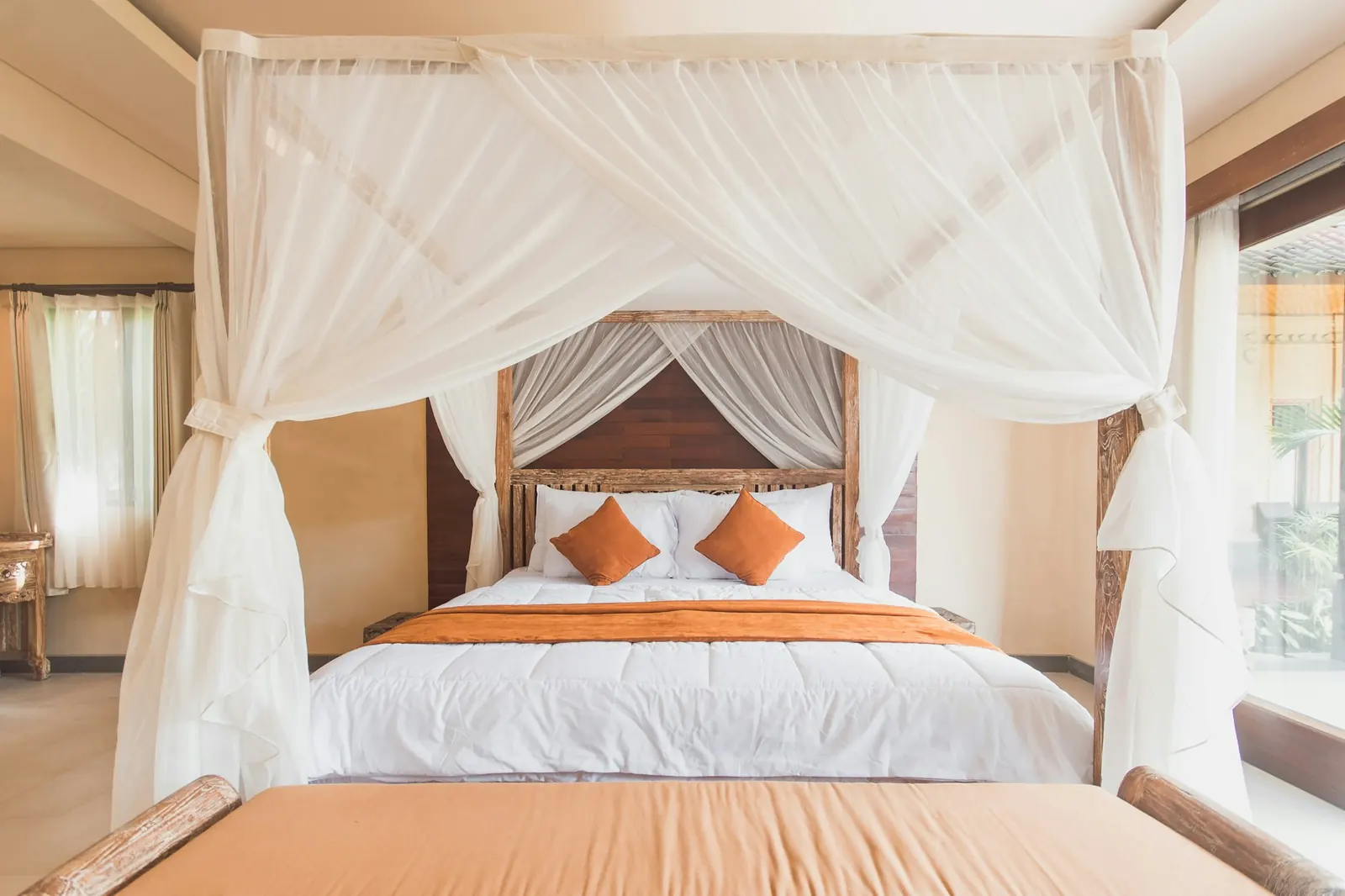 Villa bedroom with a light-wood four-poster bed draped in a white mosquito net, orange cushions, windows onto greenery