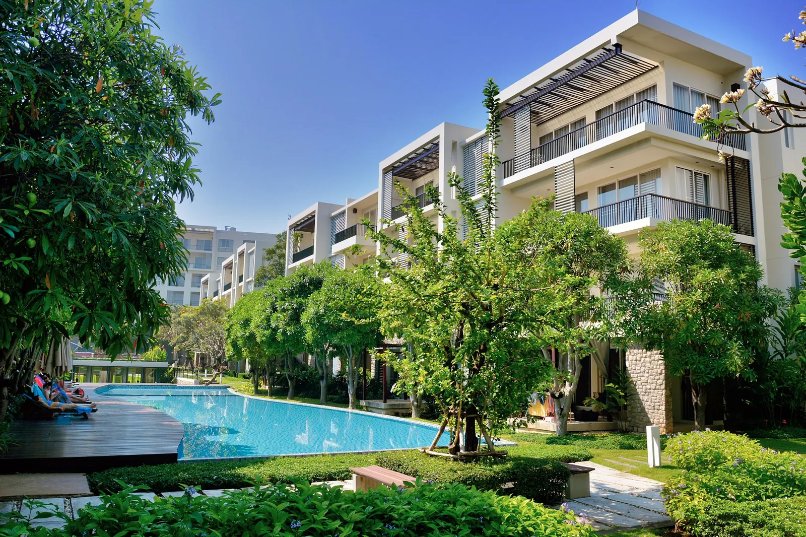 Modern four-storey residence with balconies, a long shared pool edged with lawn and tropical trees