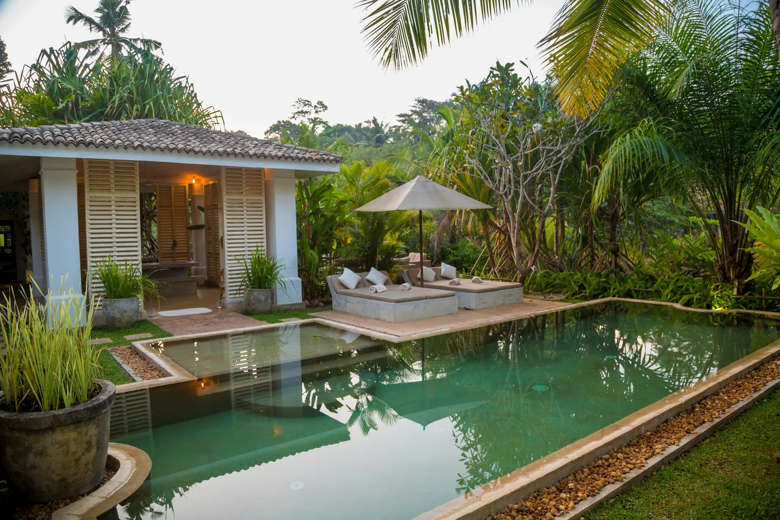 Open pavilion with wooden shutters beside a large pool, sun beds and a parasol, surrounded by palms and a tropical garden