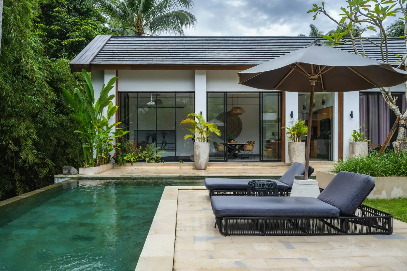 Single-storey villa with glass doors, a green-blue private pool and loungers under a parasol, in a tropical garden