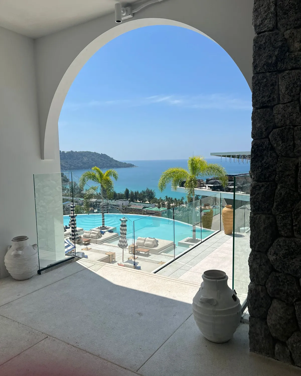 Infinity pool and the Andaman Sea seen through the white arch of a Phuket villa, palm trees and loungers by the pool
